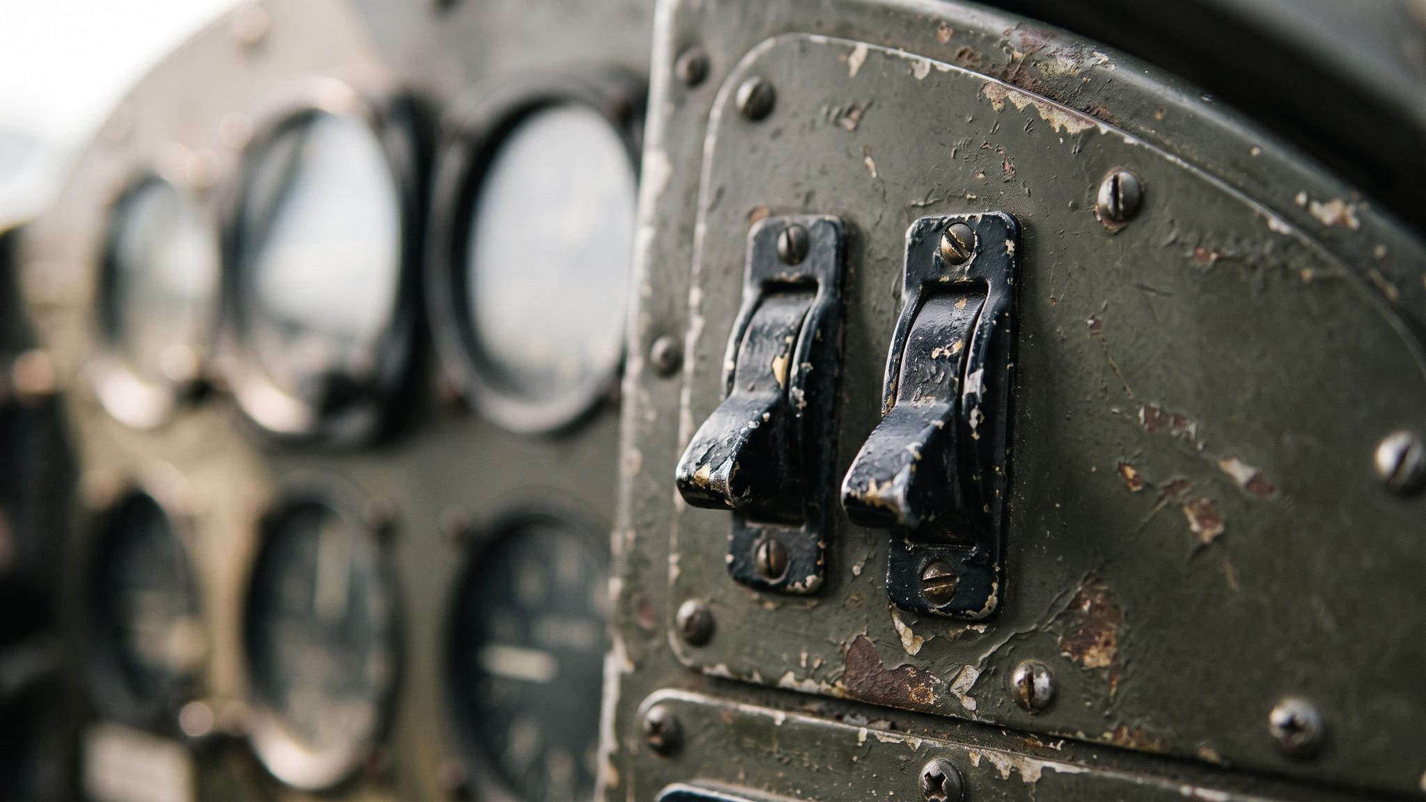 Close de um painel de avião antigo, verde-oliva e gasto: duas alavancas pretas quase idênticas, lado a lado, e os mostradores redondos desfocados ao fundo